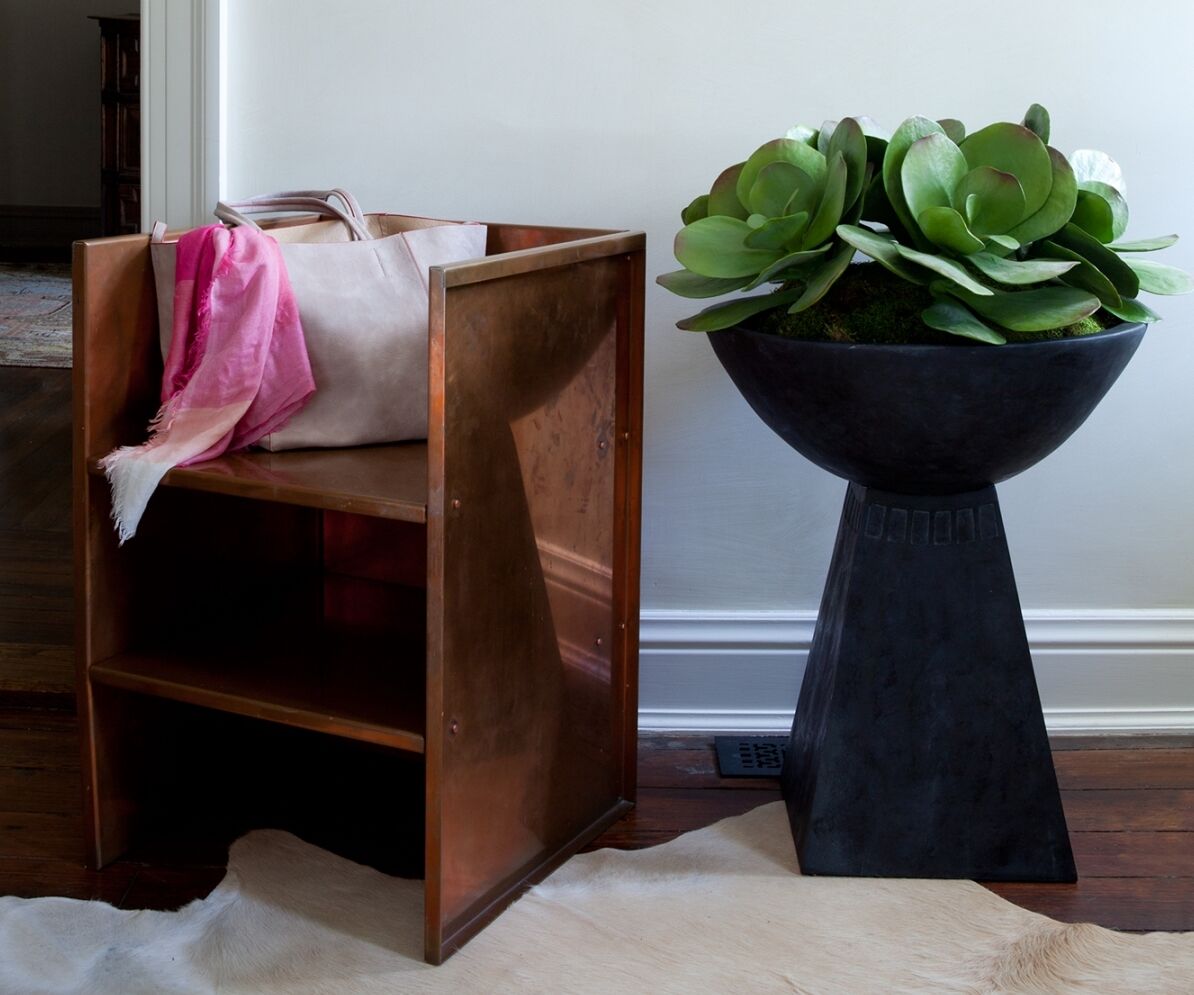 A copper-colored wooden chair with a pink and beige tote bag draped over one side sits next to a tall, black geometric plant stand holding a lush green succulent arrangement. A white fur rug lies on the wooden floor.