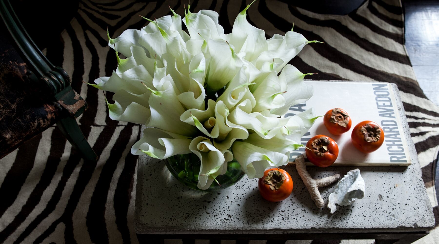 A bouquet of white calla lilies in a glass vase sits on a textured table. Nearby are four persimmons and a book titled "Richard Avedon." The table is placed on a zebra-patterned rug.