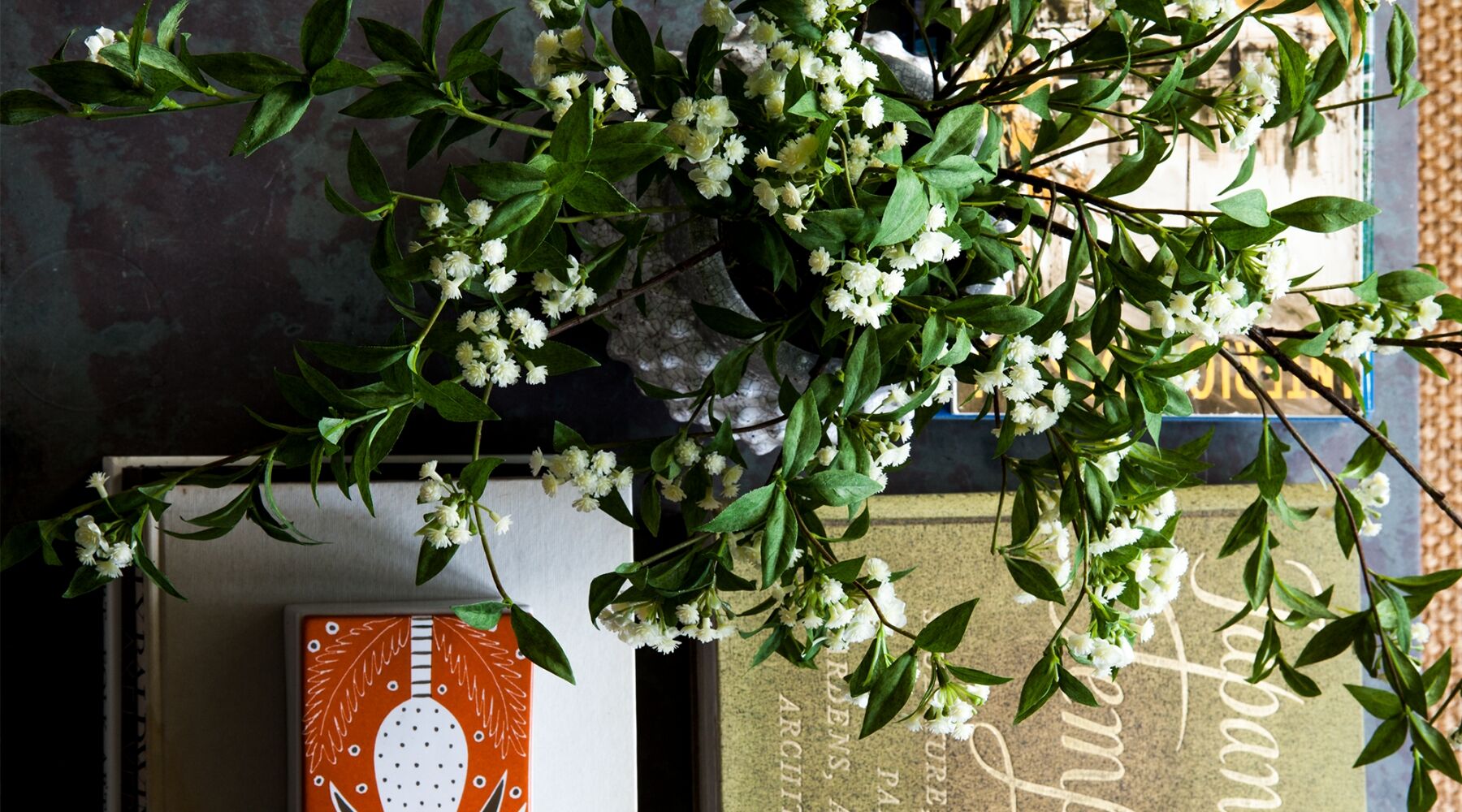 A table with a green and white floral arrangement in a vase, a hardcover book with partially visible text, and an orange and white patterned box. The setting suggests a cozy reading or living area.