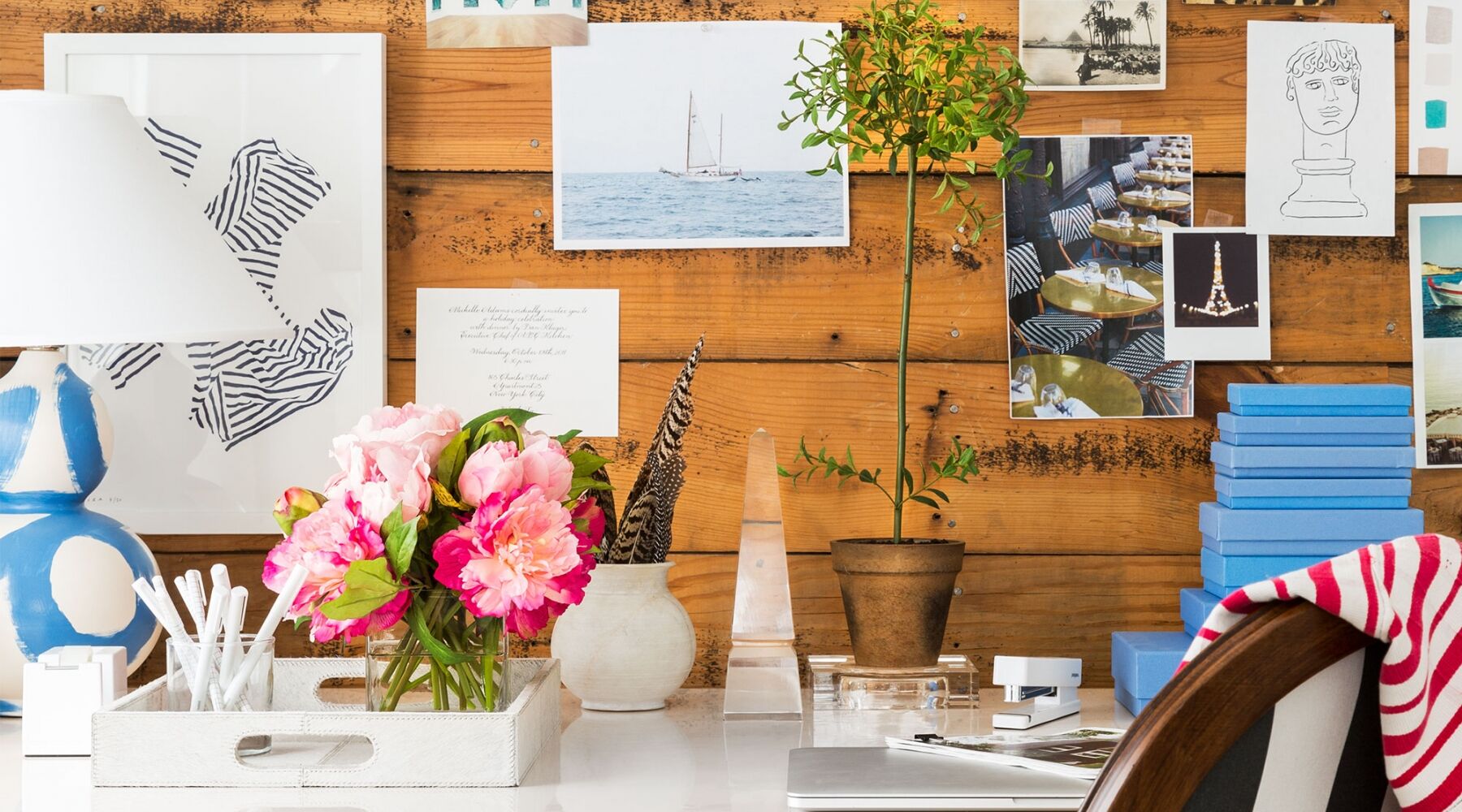 A creative workspace with a wooden backdrop covered in photos and notes. A desk holds pink flowers in a vase, a white lamp, a small potted plant, scattered art supplies, and blue boxes. A chair with a striped fabric is partially visible.