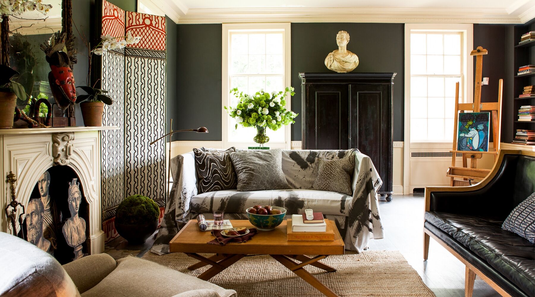 A stylish living room with dark walls, featuring a patterned couch, a wooden coffee table with books and a bowl, a bust on top of a cabinet, and an easel with a painting. Greenery decor and artistic elements add charm to the space.