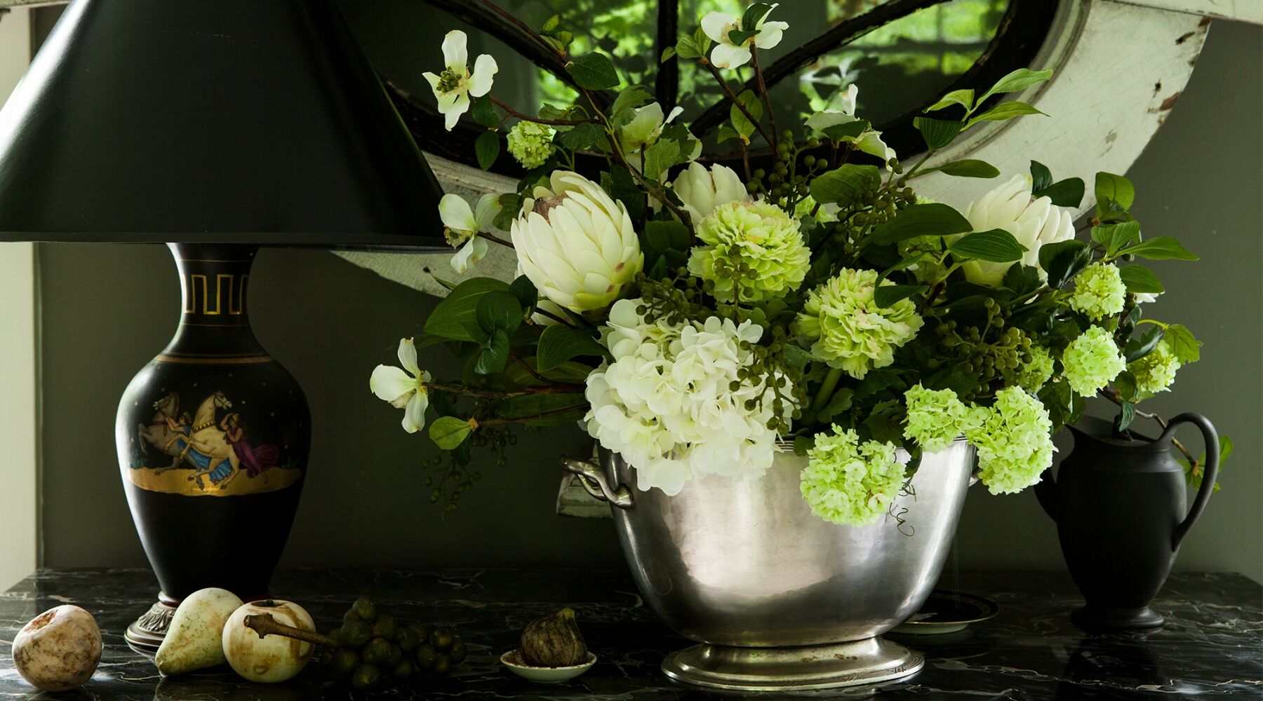 A large silver vase filled with white and green flowers sits on a dark marble table. To the left, there's a black and gold lamp, and to the right, a small black pitcher. Two pears and a shell decorate the table in front.
