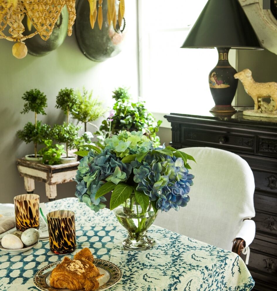 A cozy dining area with a table set with a blue-patterned cloth, a glass vase of blue hydrangeas, and a plate of pastries. In the background are potted plants, a decorative lamp, and a figurine on a black dresser. Natural light filters in.