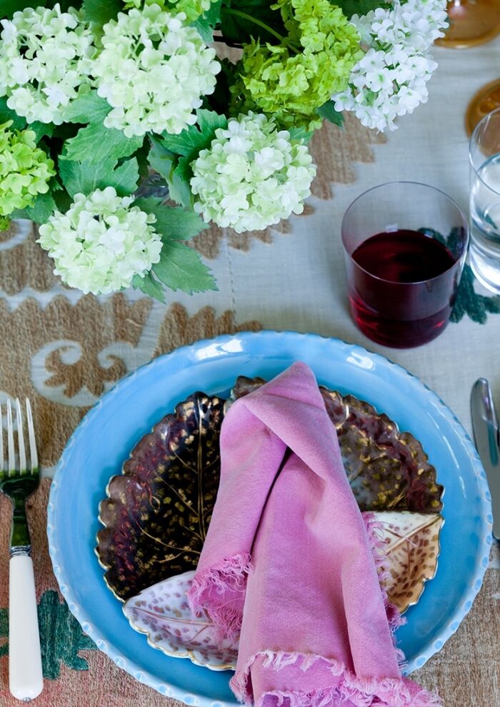 A table setting with a blue plate, layered dark decorative plates, and a pink cloth napkin. Beside it are a fork, knife, glass of red liquid, and water. Green and white flowers are arranged above on a patterned tablecloth.