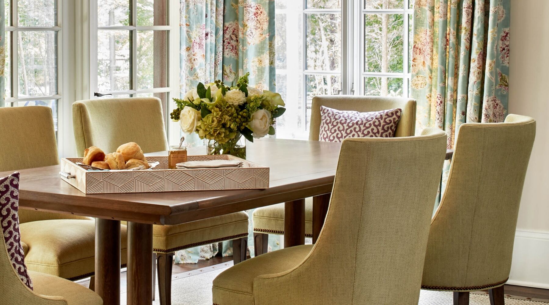 A dining room with a wooden table and six green upholstered chairs. On the table, there's a decorative tray with bread rolls and a vase of white flowers. The room is filled with natural light from large windows with floral curtains.