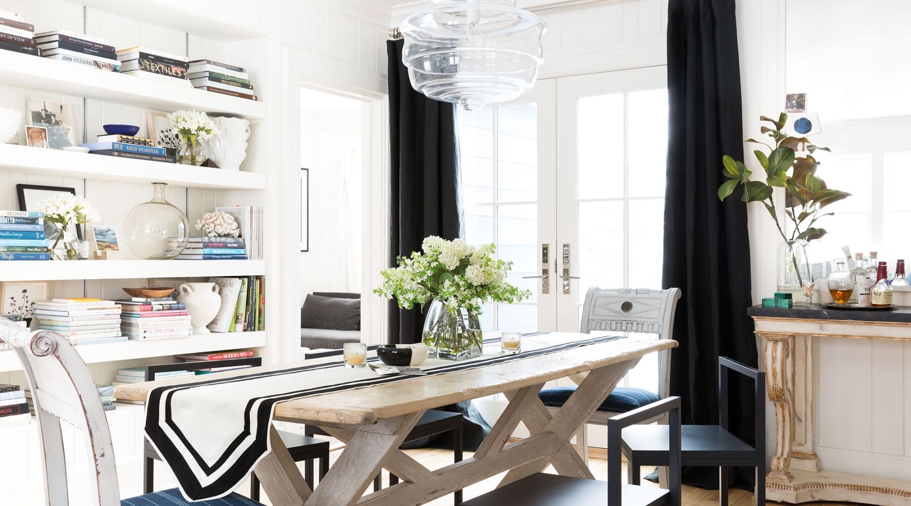A bright dining room with a wooden table, black chairs, and a vase of flowers. The table has a black and white runner. Books and decor fill the shelves. A clear pendant light hangs above. Black curtains flank large windows in the background.