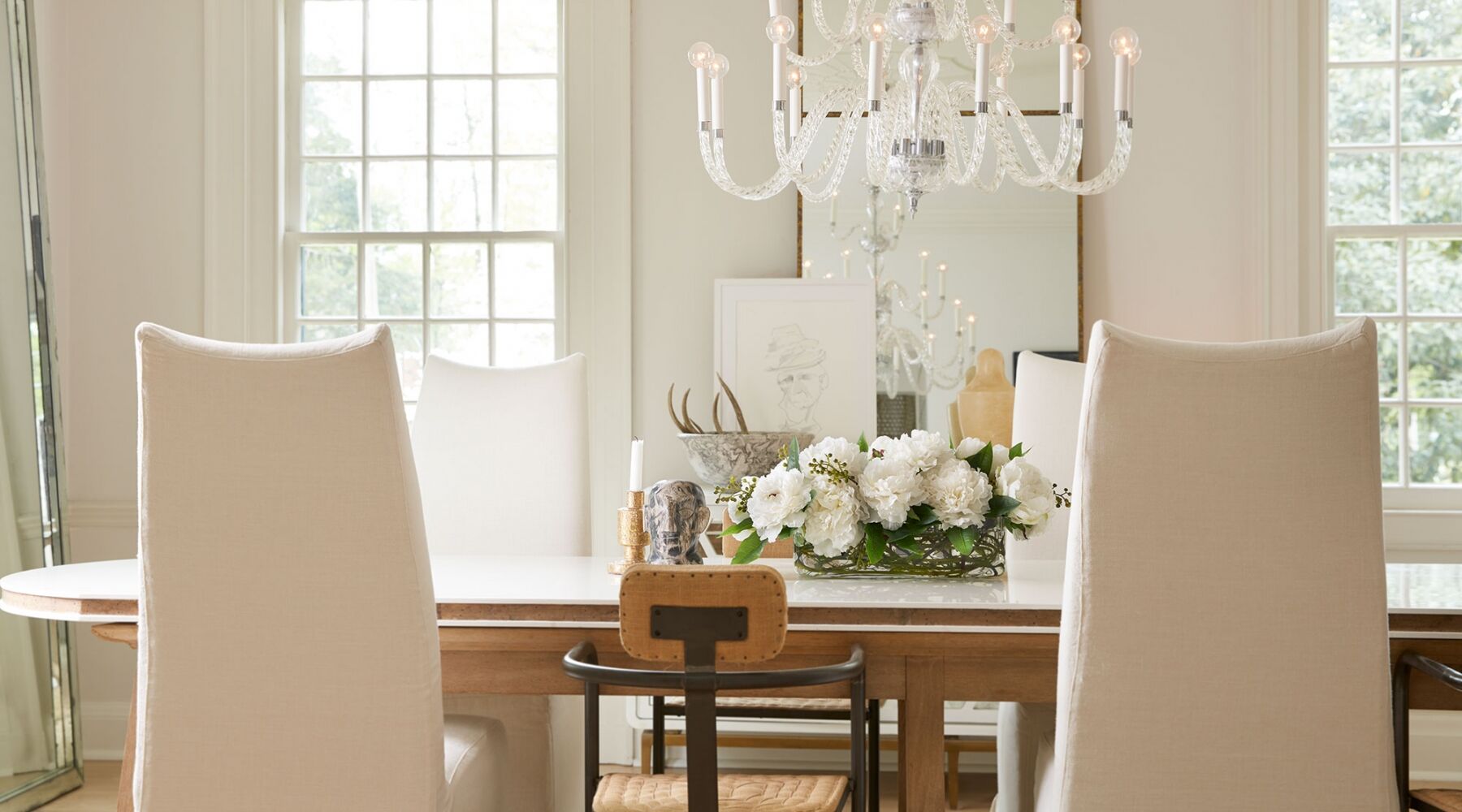 A bright dining room with a wooden table, white upholstered chairs, and a crystal chandelier. A centerpiece of white flowers sits on the table. Large windows in the background let in natural light.