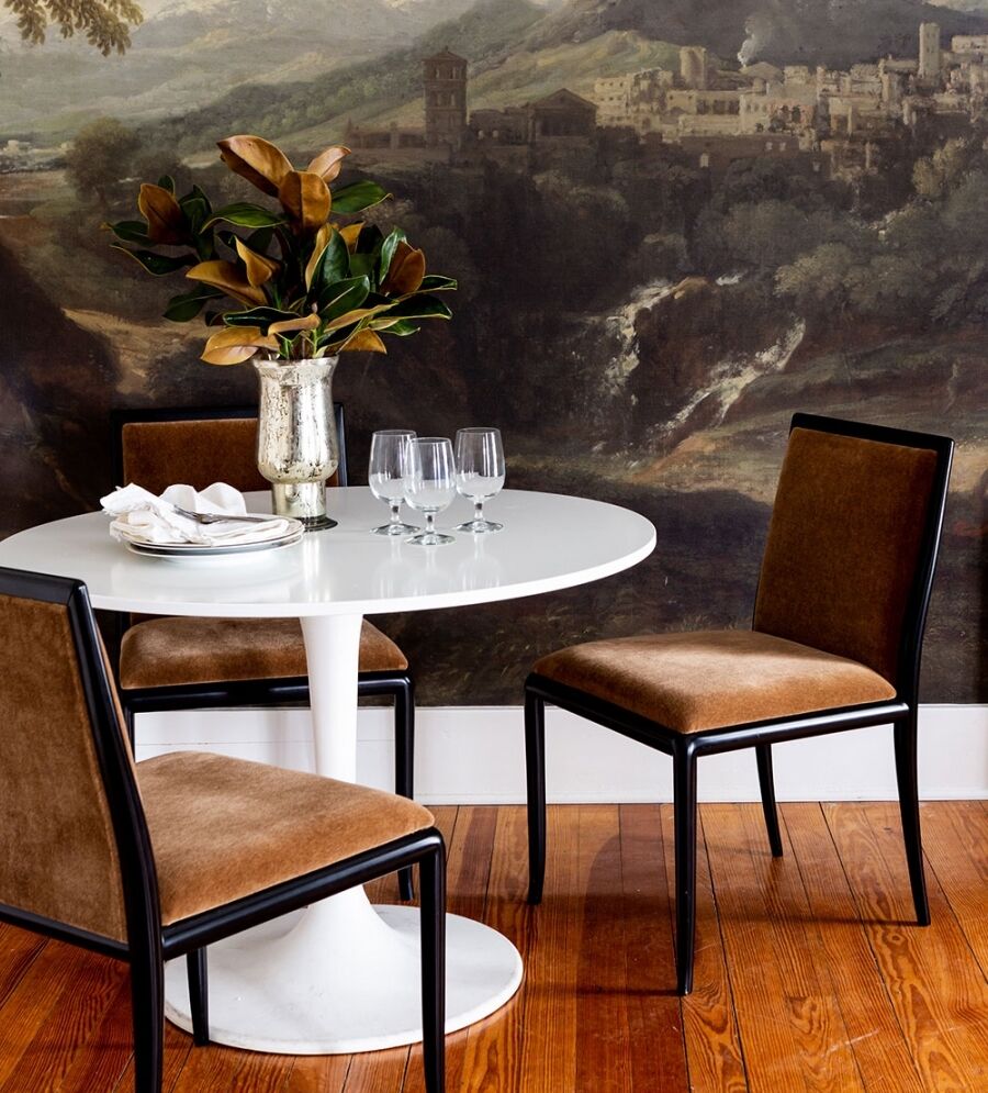A round white table with three brown chairs is set with three wine glasses and a small stack of plates. A vase with leaves sits on the table. The background features a large painting of a landscape with mountains and a village.