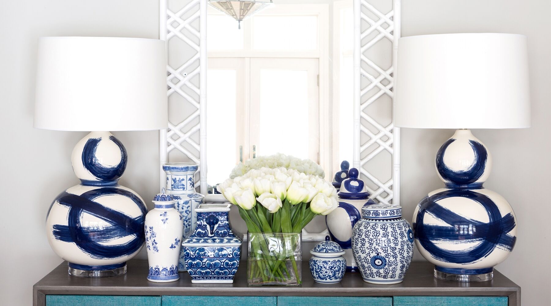 A decorative arrangement featuring two white lamps with navy blue brushstroke designs, various blue and white patterned vases, and a transparent vase with white flowers on a teal console under a lattice-patterned mirror.