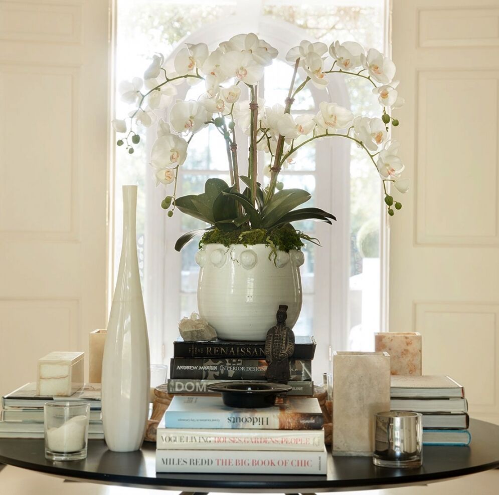 A round table with a white orchid in a pot as the centerpiece. The table is adorned with various books stacked in piles, two white vases, small candles, and decorative items. Soft natural light filters in through a window in the background.