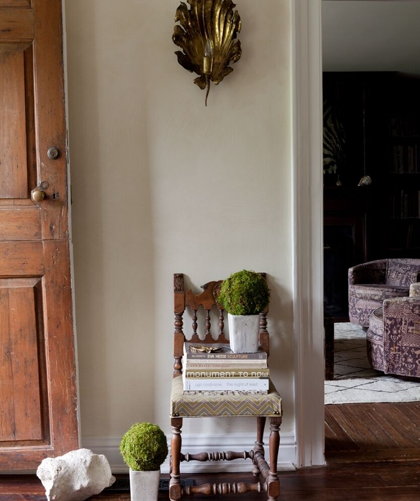 A rustic wooden chair with patterned cushions holds two small potted plants and a stack of books. A vintage sconce is mounted on the beige wall above. A wooden door is slightly ajar, leading to a warmly lit room with a purple patterned sofa.