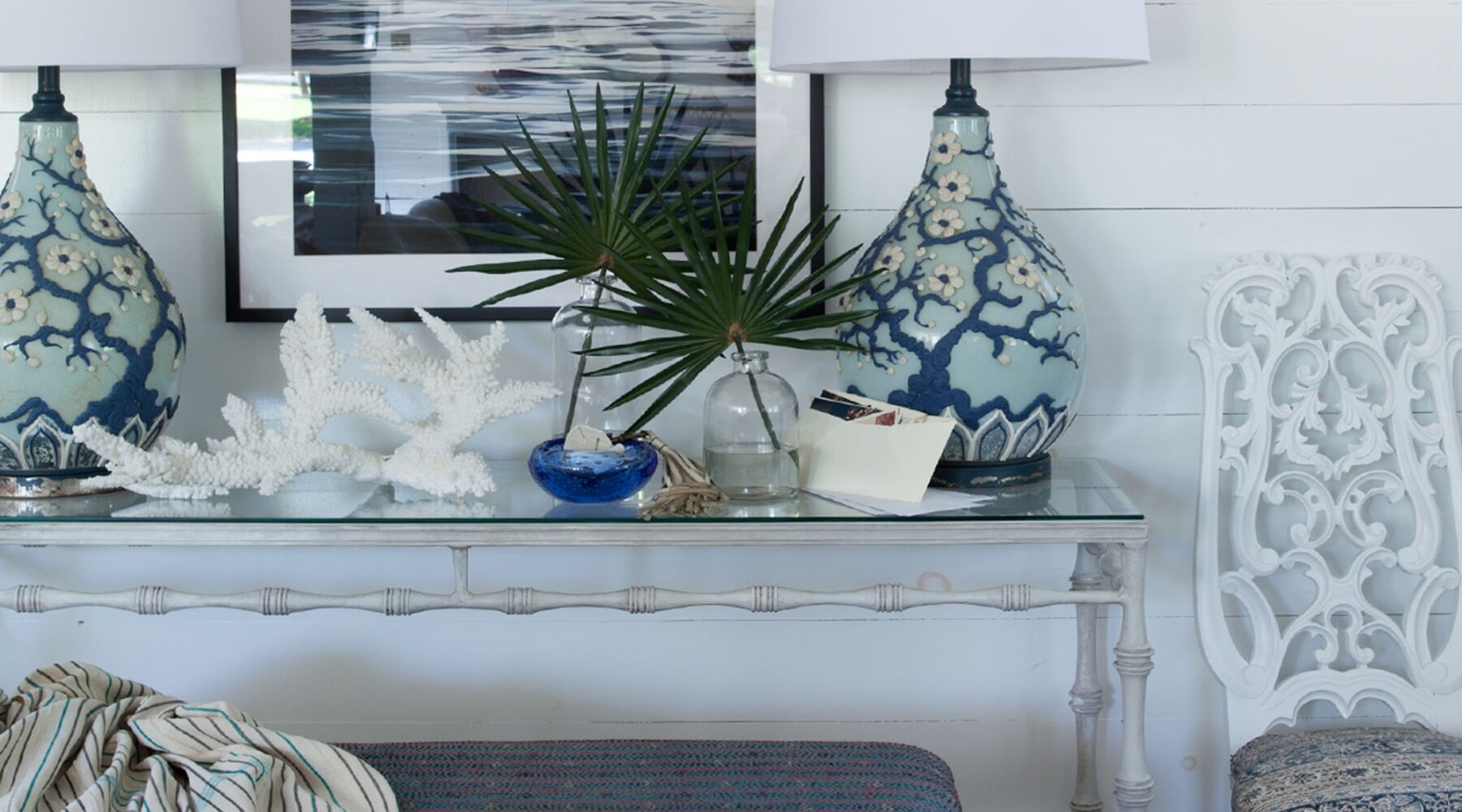 A glass-top table with two large, decorative blue lamps, a white coral sculpture, and a small potted plant. A framed picture hangs on the wall behind, and a white carved chair is partially visible on the right.