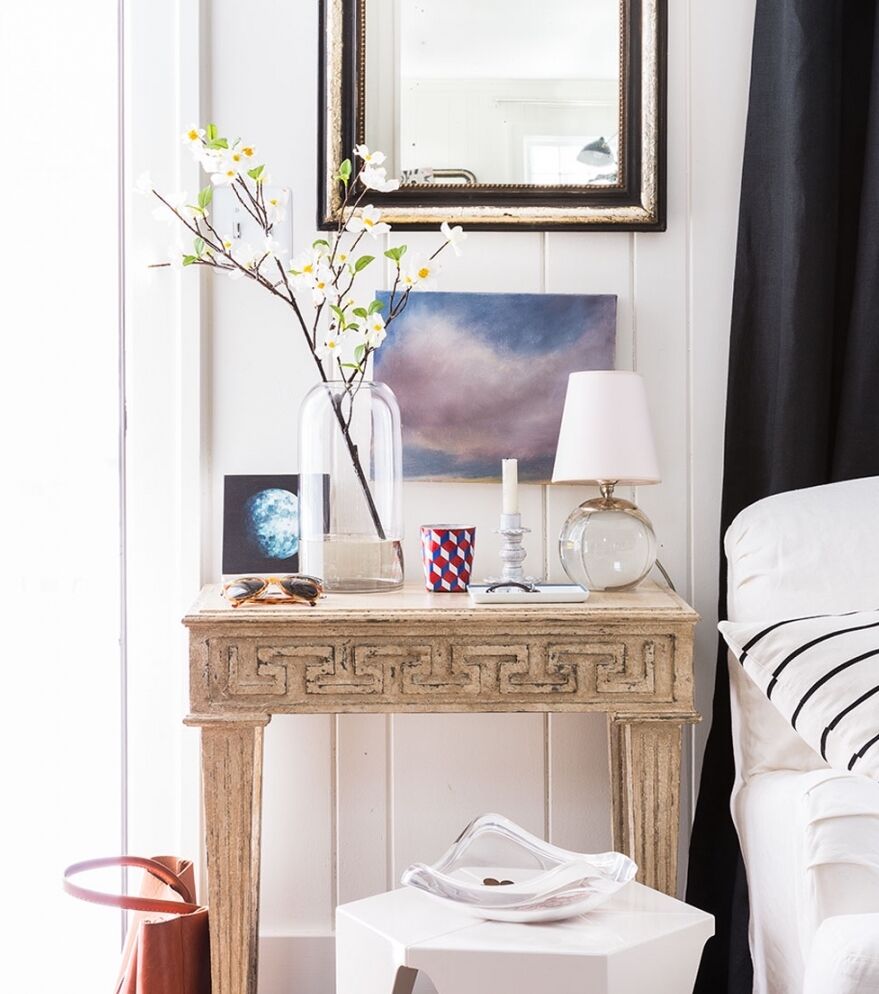 A small wooden table with a Greek key design holds a vase of flowers, a lamp, framed art, and a candle. A mirror hangs above. Next to the table is a white sofa and a contemporary side table with a decorative object. A brown bag is on the floor nearby.