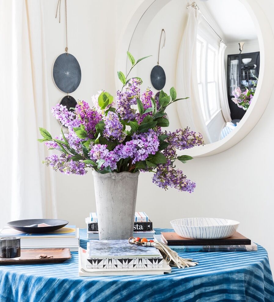 A table with a blue striped cloth features a vase of purple flowers, surrounded by stacked books, a bowl, and a cup. A round mirror reflects the room, and two circular wall decorations hang nearby.