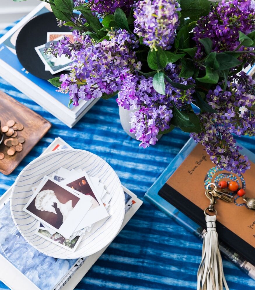 A table decorated with a vase of purple flowers, a stack of books, a dish holding several vintage photos, a set of keys with colorful keychains, and a small pile of coins. The tablecloth is a striped blue fabric.