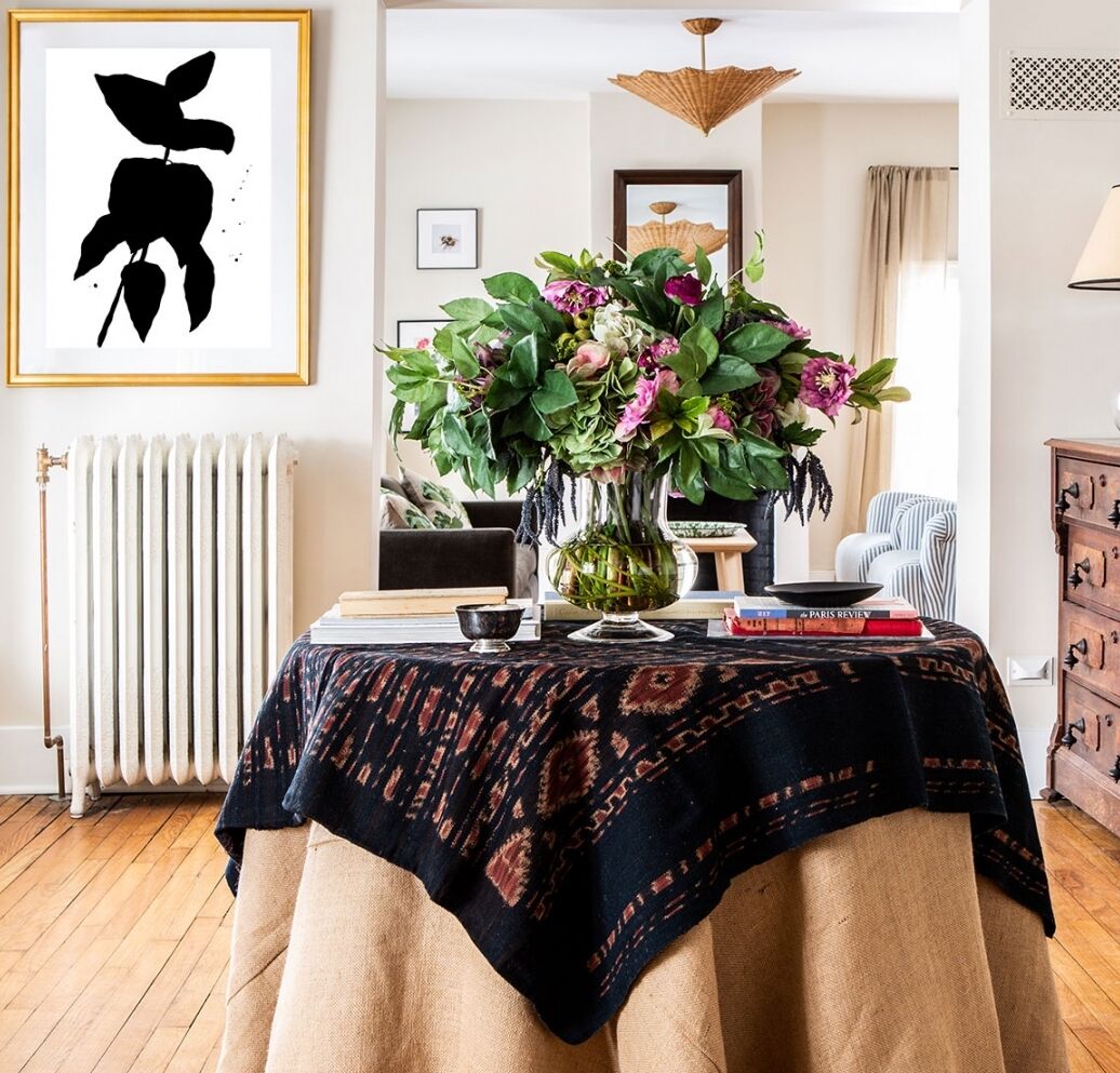 A cozy living room with a round table covered by a patterned cloth. On the table, there's a vase of flowers, books, and a coffee cup. A monochrome plant print hangs on the wall. A radiator, armchair, and wooden dresser complete the space.