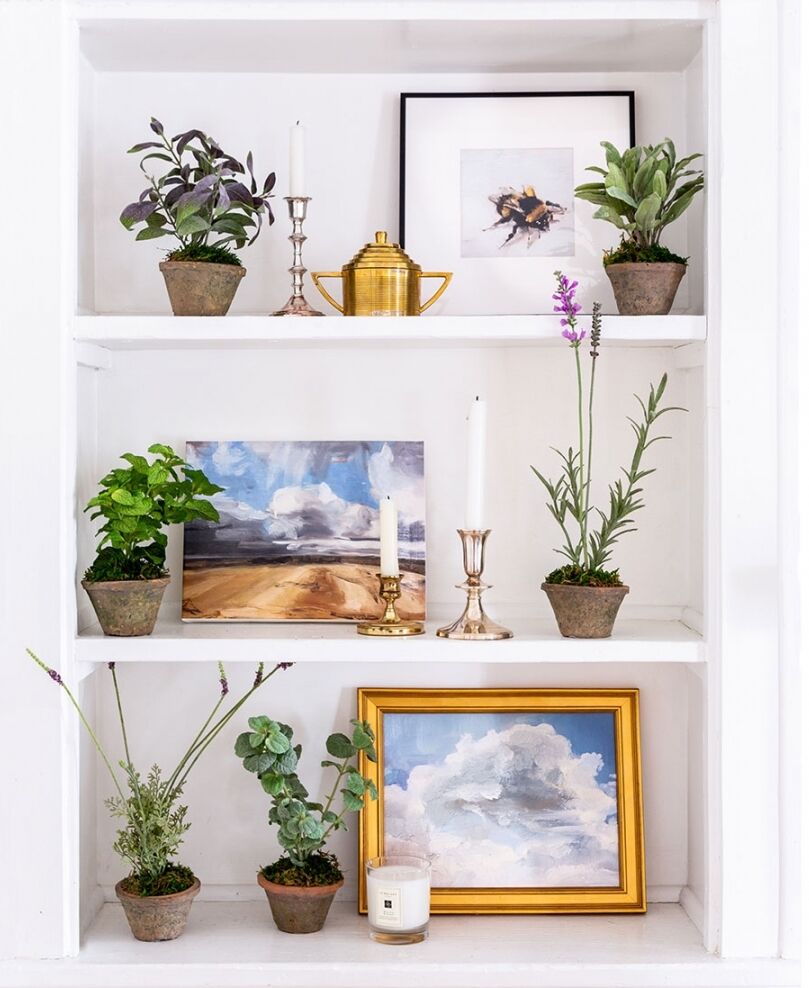 A white shelf displaying potted plants, framed art, and candles. The arrangement includes green foliage and small flowering plants alongside artworks of landscapes and a bee, creating a cozy and artistic decor.