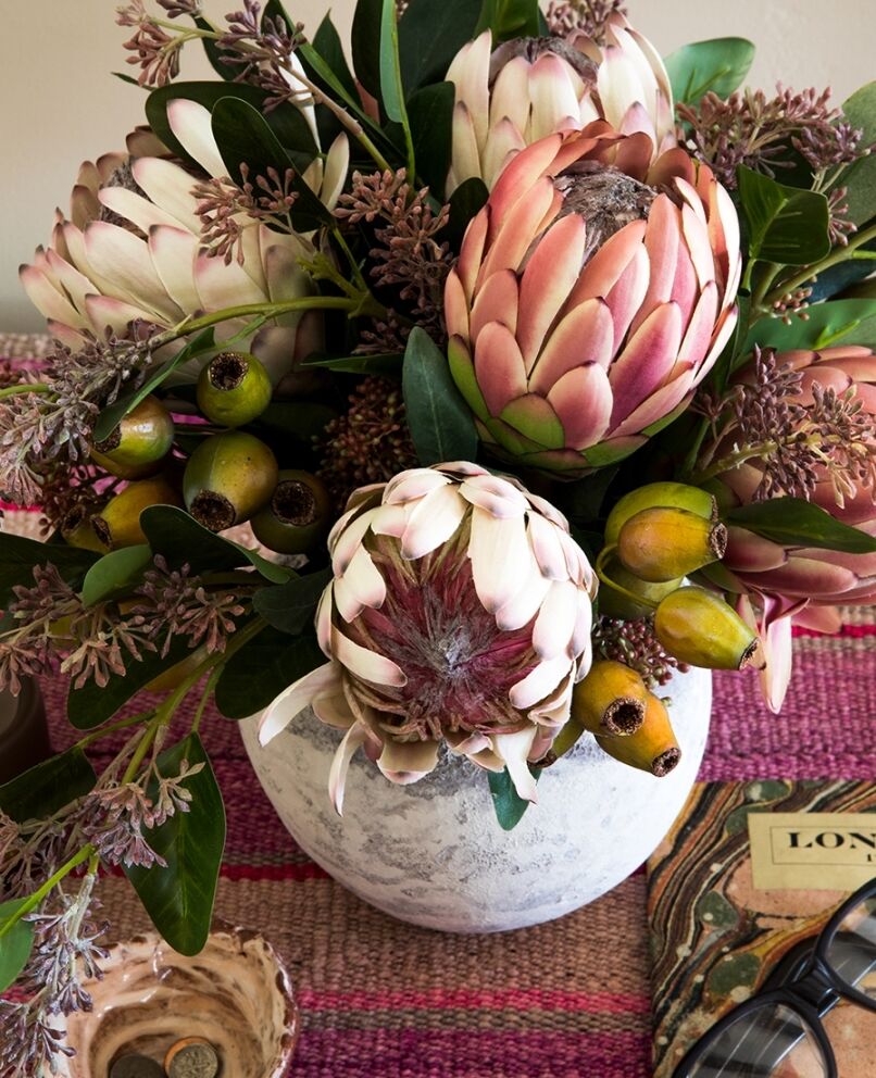 A textured white vase holds a vibrant floral arrangement featuring large pink protea flowers, green leaves, and small clusters of pinkish buds. The vase is placed on a woven pink mat, next to a book and a pair of glasses.