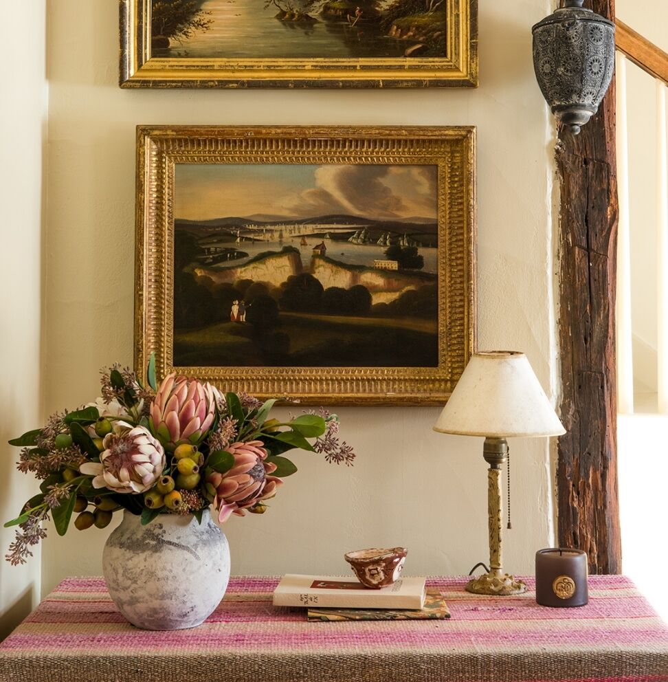 A table with a pink and beige cloth holds a vase of flowers, a book, a decorative cup, a small lamp, and a candle. A framed landscape painting hangs on the wall above, with a traditional lamp fixed on the wall to the right.
