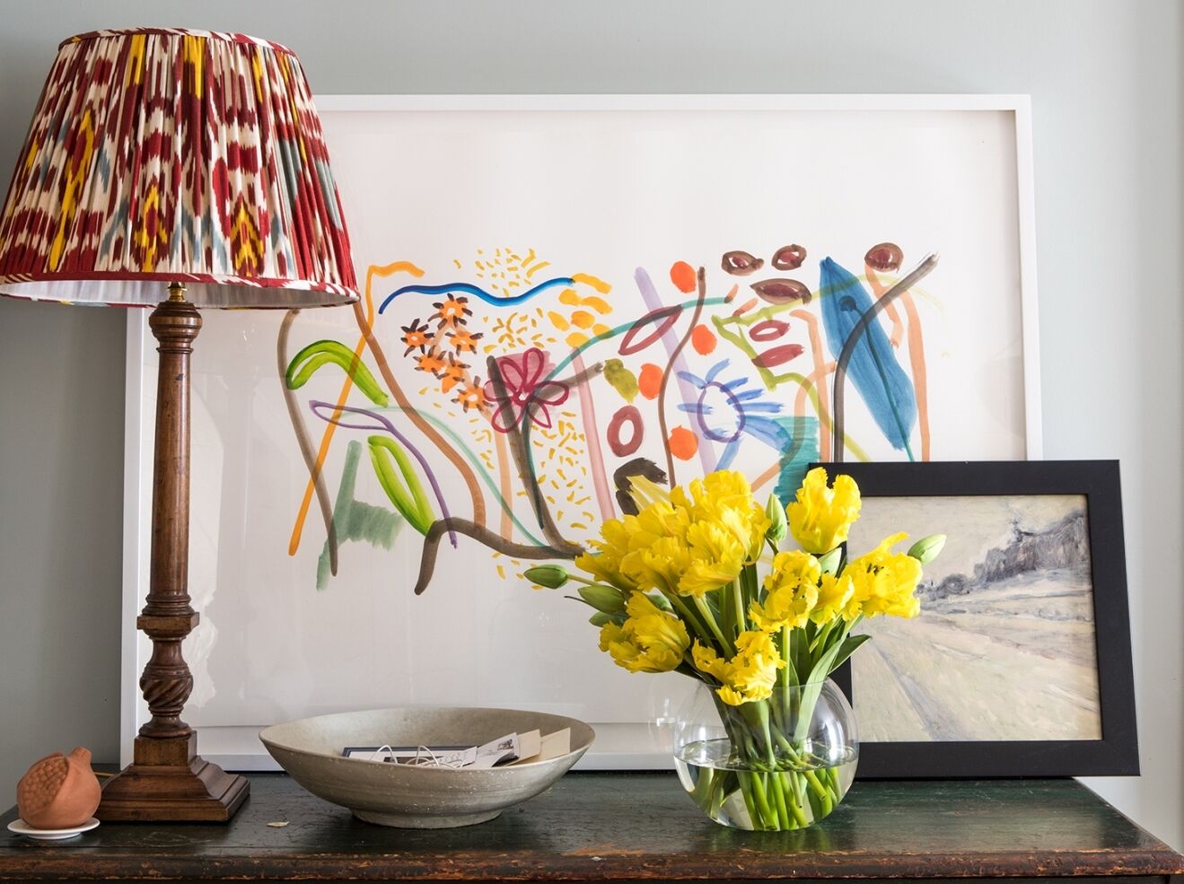 A colorful abstract painting hangs above a wooden shelf, accompanied by a tall lamp with a patterned shade. A clear vase holds bright yellow flowers beside a small framed landscape painting and a bowl containing mail.