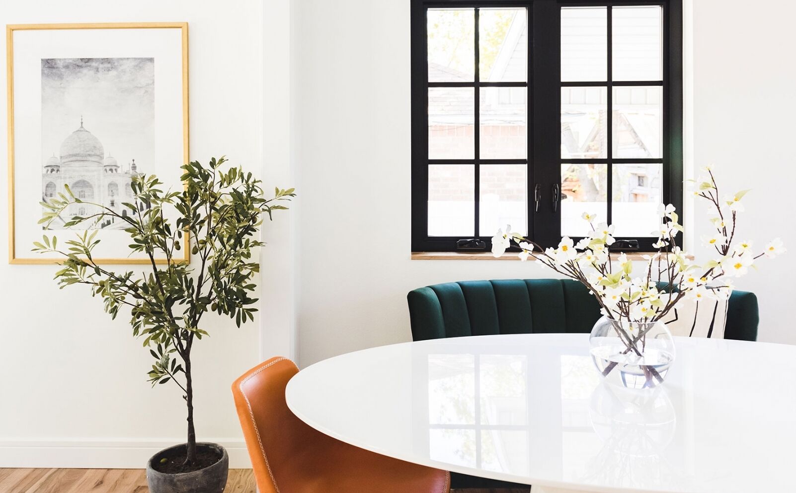 A bright, modern dining room features a round white table with a vase of white flowers, orange and green chairs, a potted plant, a framed black-and-white artwork on the wall, and black-framed windows with a view of another building.