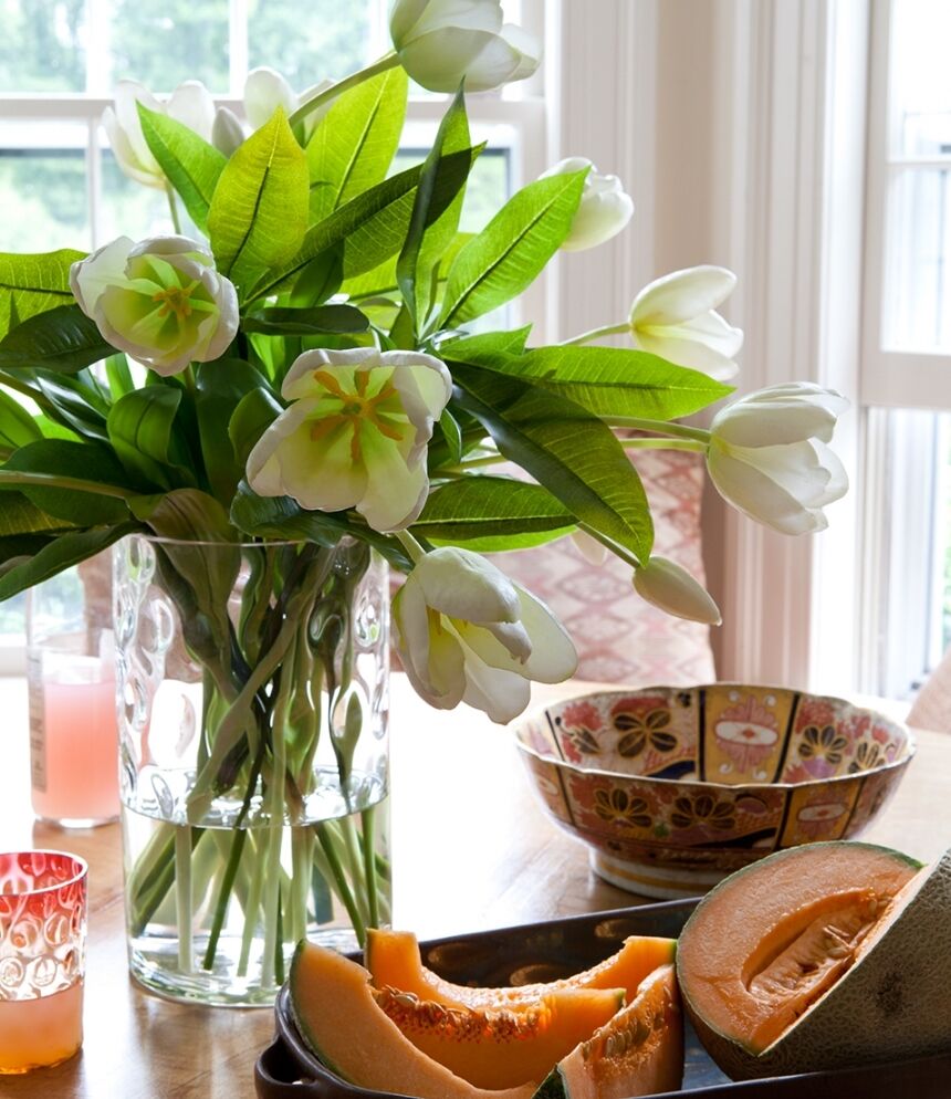 A bouquet of white tulips in a clear vase on a table with sliced melon, a decorative bowl, and two patterned glasses. Sunlight filters through a window in the background.