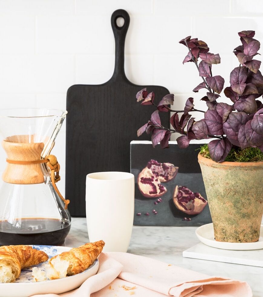 A cozy kitchen scene with a coffee maker, a white mug, a croissant on a plate, a potted plant, and a wall art featuring pomegranates. A wooden cutting board adds a rustic touch.
