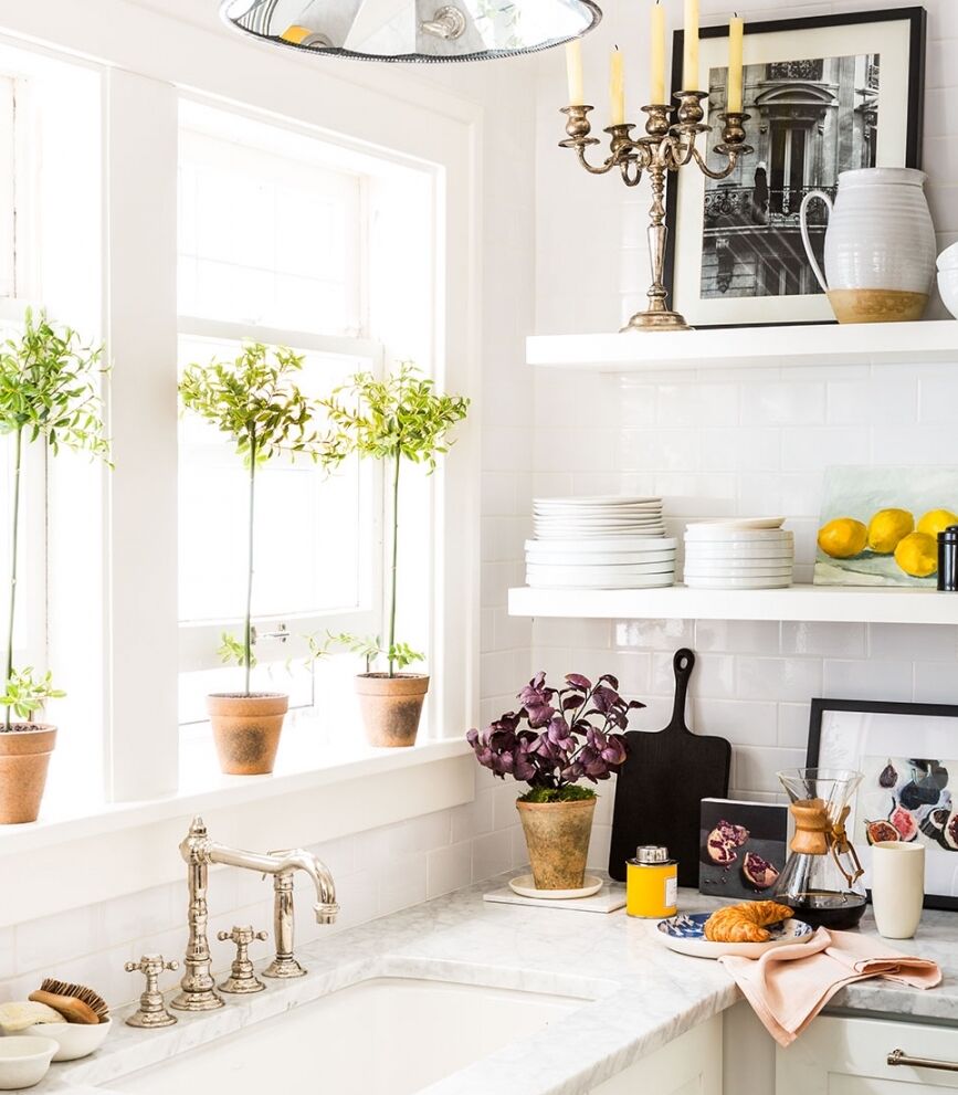 A bright kitchen with a marble countertop, white shelves, and a window with potted plants. The shelves display dishes, lemons, and decorative items. A croissant and a juice glass are on the counter, creating a cozy, inviting atmosphere.