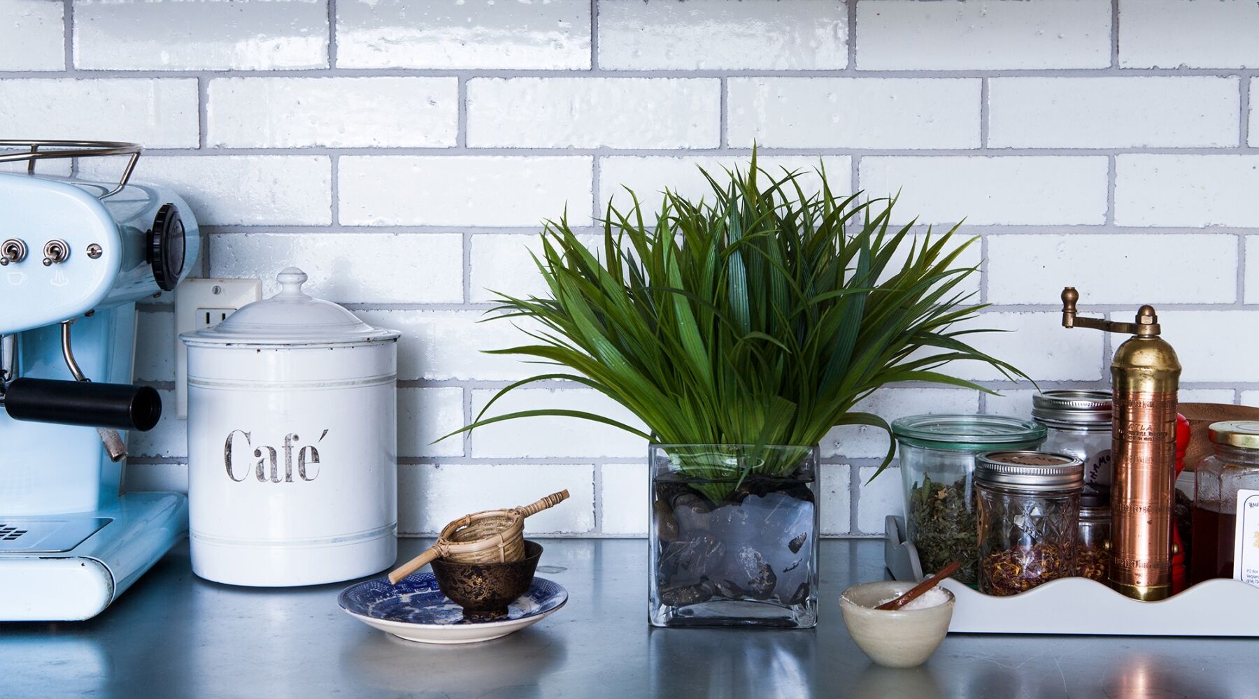 A kitchen counter with a light blue espresso machine, a white container labeled "Café," a potted green plant, and an assortment of jars with spices and a vintage coffee grinder on a white tray. White subway tile backsplash in the background.