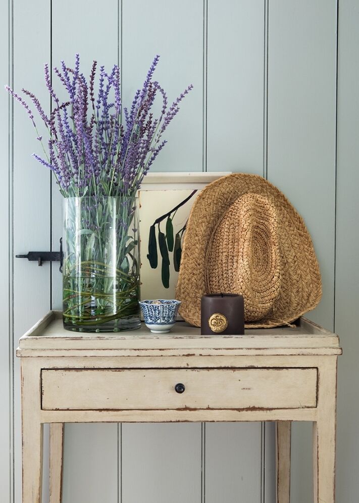 A small table with a glass vase of lavender, a straw hat, a small blue and white teacup, and a brown candle. A simple wall with vertical panels is in the background.