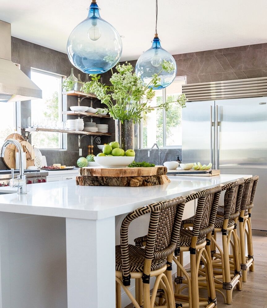 Modern kitchen with a large island featuring woven bar stools and a white countertop. A tree slice serves as a centerpiece with green apples. Hanging glass pendant lights illuminate the space, with stainless steel appliances in the background.