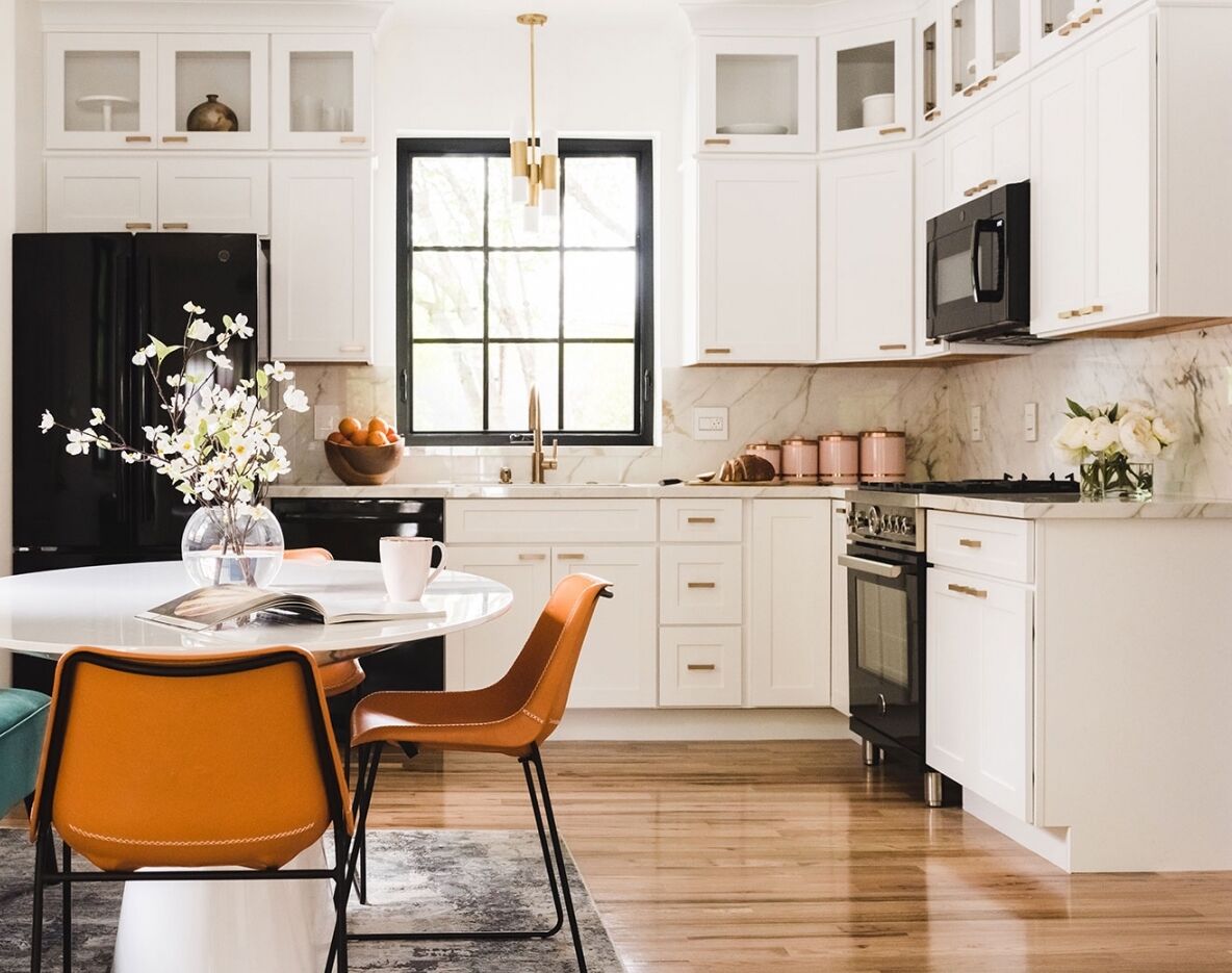 Modern kitchen with white cabinets, a black fridge, and oven. A round table with orange chairs is in the foreground, adorned with a vase of flowers. The room features wooden flooring, a window, and elegant gold accents.