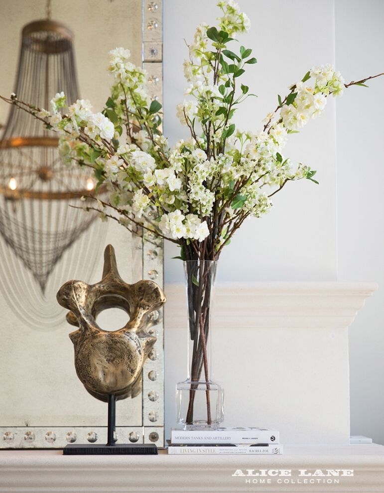 A decorative table arrangement featuring a tall glass vase with white flowering branches and a sculptural golden art piece on a stand. Nearby, books are stacked on the table, with a large mirror and chandelier in the background.