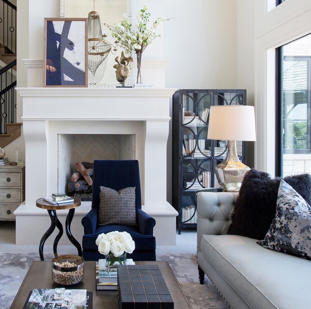A stylish living room with a white fireplace, navy armchair, and light gray sofa adorned with dark pillows. A modern side table holds books and a vase of white flowers. A tall lamp and bookshelf add warmth. Decorative art and vases embellish the mantel.