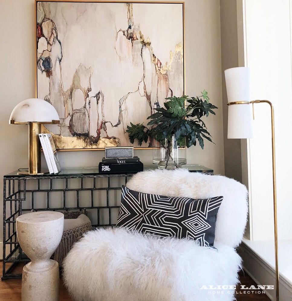 A cozy seating area featuring a fluffy white chair with a black geometric pillow, a small round side table, and a glass console table. A large abstract painting hangs on the wall above, with a tall plant and a modern floor lamp nearby.