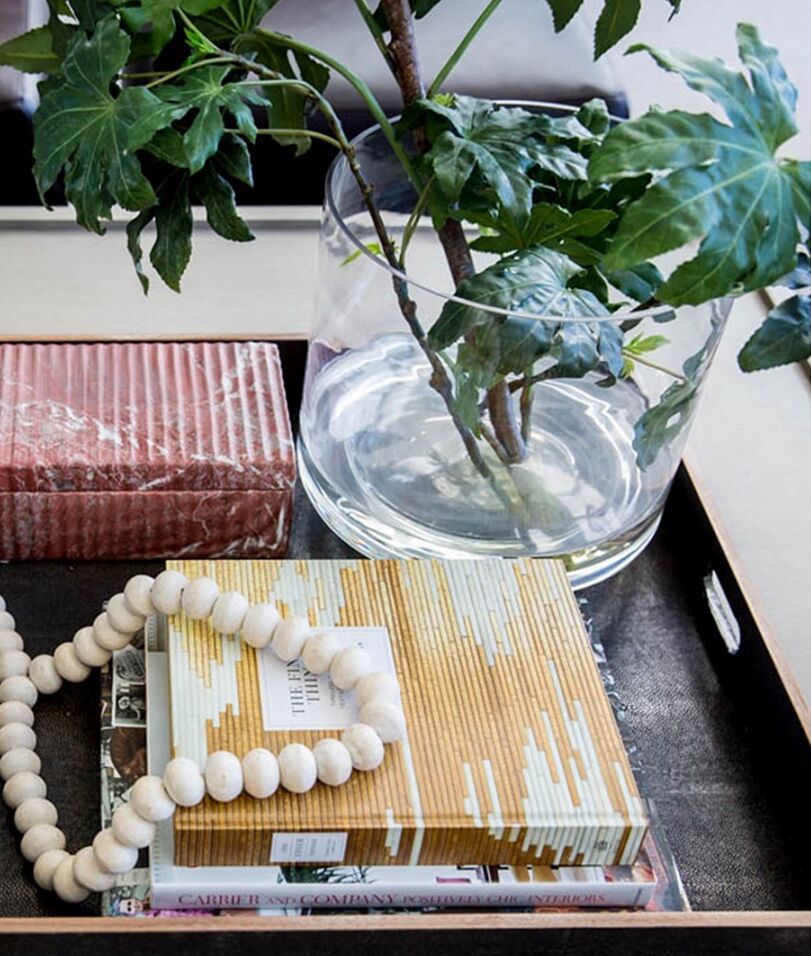 A decorative tray on a table holds a clear vase with leafy branches, a glass brick, books with artistic covers, and a strand of wooden beads. The arrangement creates a sophisticated and natural aesthetic.