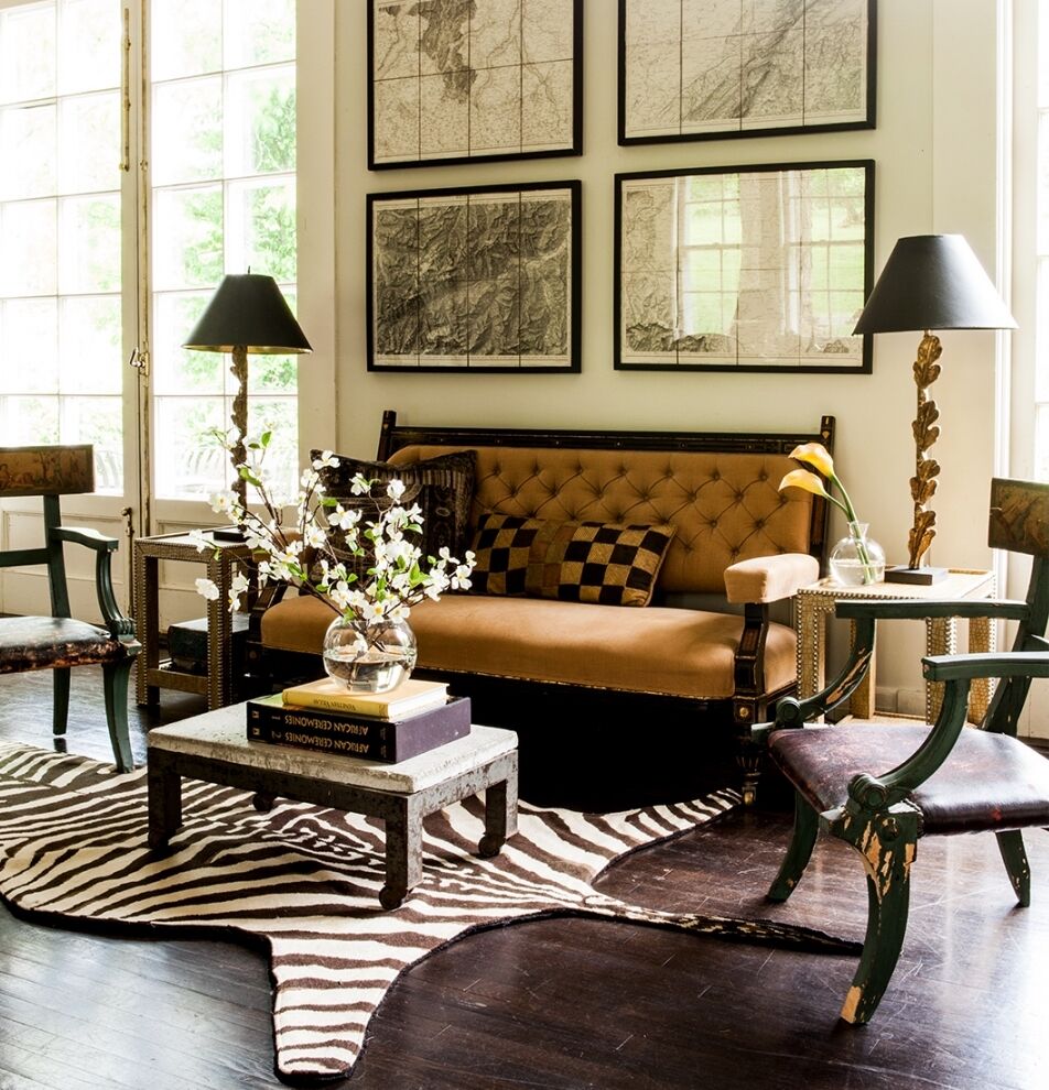 A cozy living room with a brown tufted sofa, decorative pillows, and a zebra-patterned rug. The room features dark wood furniture, two black lamps with gold accents, and framed art on the wall, complemented by large windows.