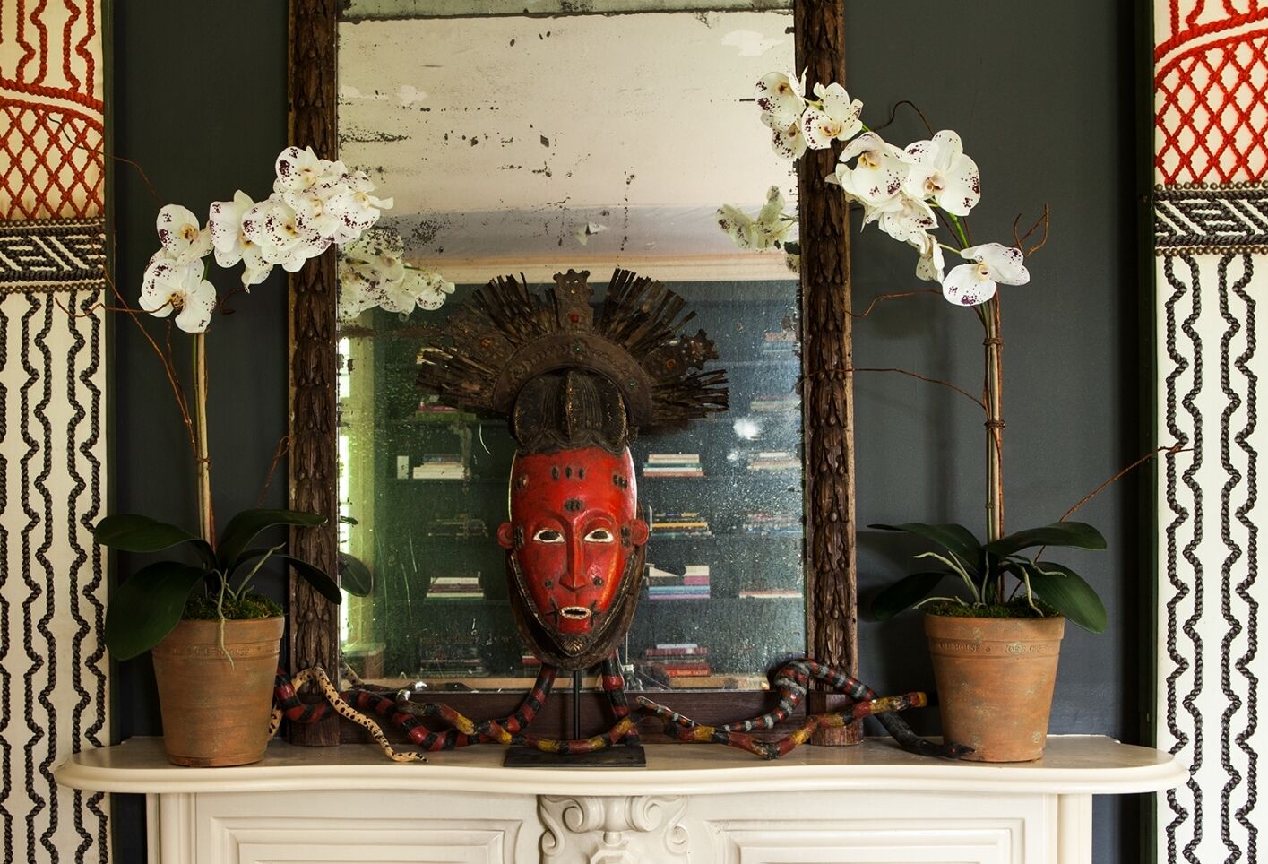 A decorative red tribal mask with dark crown-like adornments is displayed on a mantel. Flanked by potted orchids with white blooms, it's reflected in a large, rustic mirror. A colorful fabric is draped across the surface.