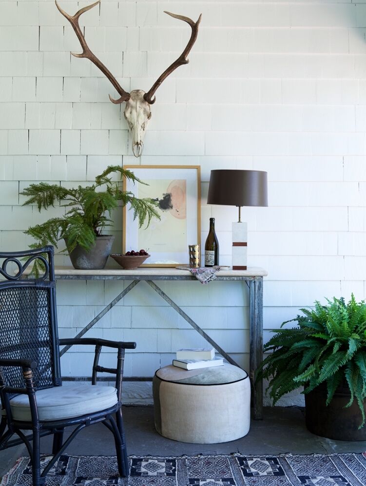 A rustic porch features a wall-mounted antler display, a wooden table with potted plants, framed artwork, a lamp, a wine bottle, and glass. A woven chair, a cushioned stool with books, and a patterned rug complete the cozy setting.