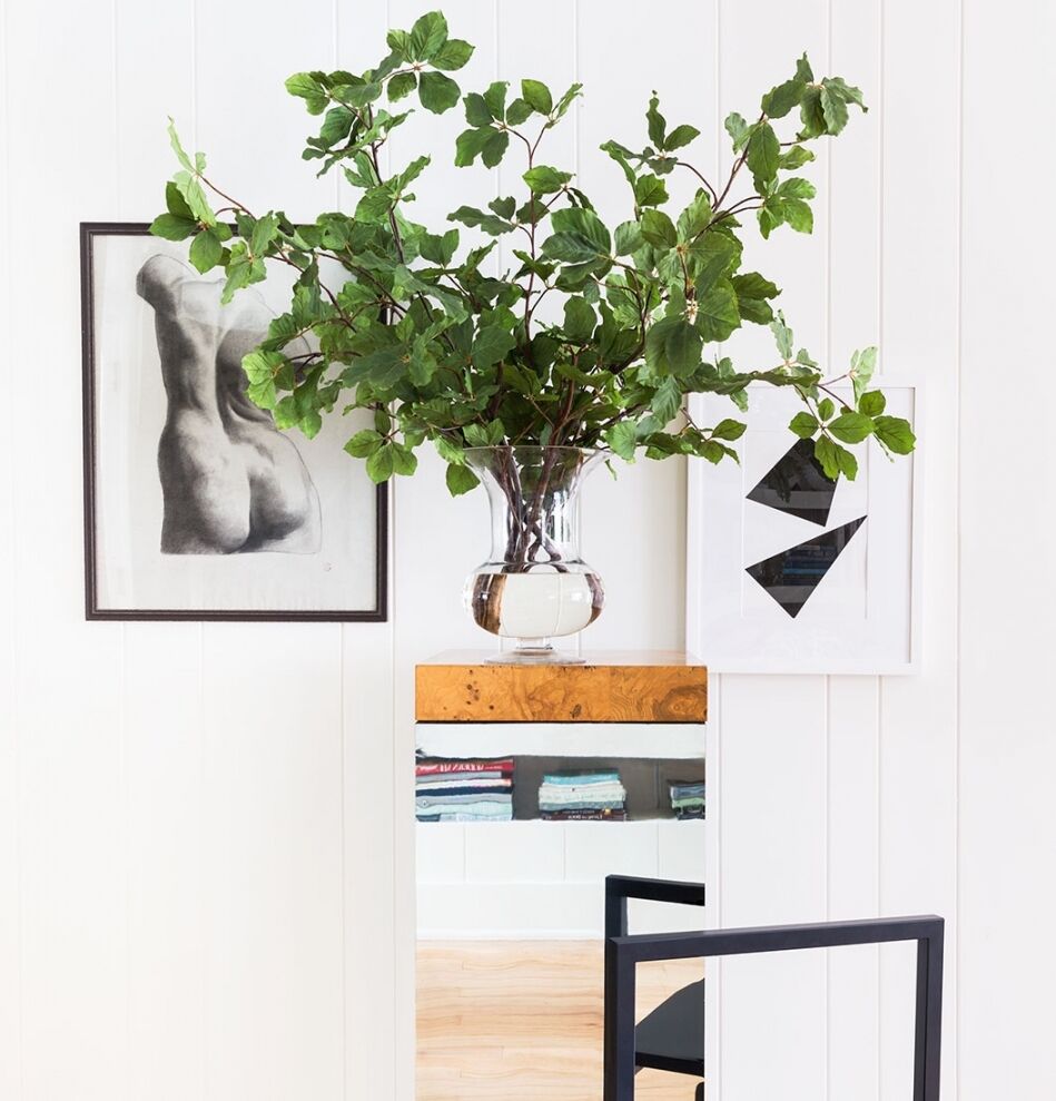 A minimalist interior with a clear vase holding green leafy branches on a mirrored table. The wall features a black-and-white abstract art piece and a framed sketch of a human torso. A black chair is partially visible in the foreground.