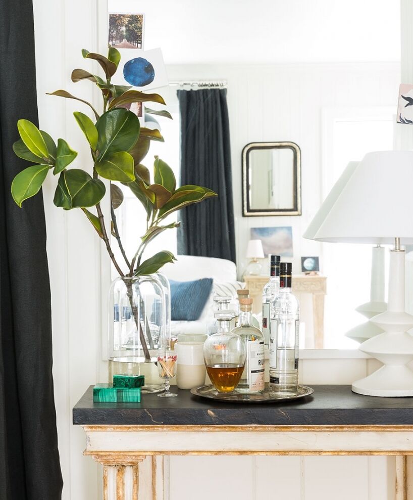 A stylish room features a table with a potted plant, various bottles, and a lamp. A mirror reflects a bed and additional decor, adding depth to the bright, modern space. A dark curtain contrasts with the light walls and furniture.