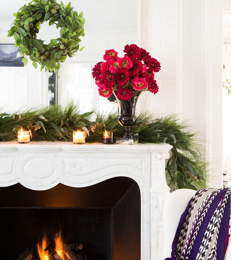 A cozy fireplace scene features a marble mantel adorned with evergreen garland, red flowers in a black vase, and lit candles. A green wreath hangs above, and a purple-striped blanket drapes over a nearby chair.