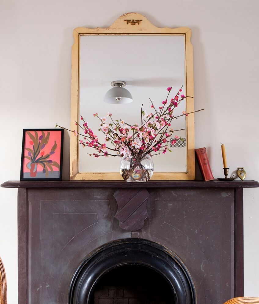 A vintage mirror with a gold frame is mounted above a dark wooden fireplace mantel. On the mantel are a framed artwork, a vase with pink flowers, a candlestick, a small sculpture, and a red book. A ceiling light is reflected in the mirror.