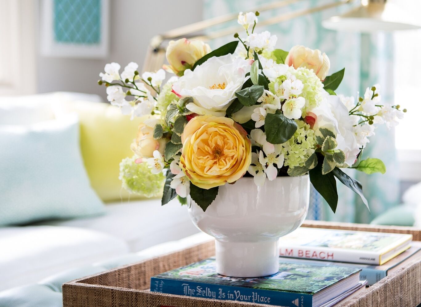 A white ceramic vase holds a bouquet of yellow and white flowers with green leaves, placed on a table with books. The background features a softly lit living room with a white sofa and pastel-colored cushions.