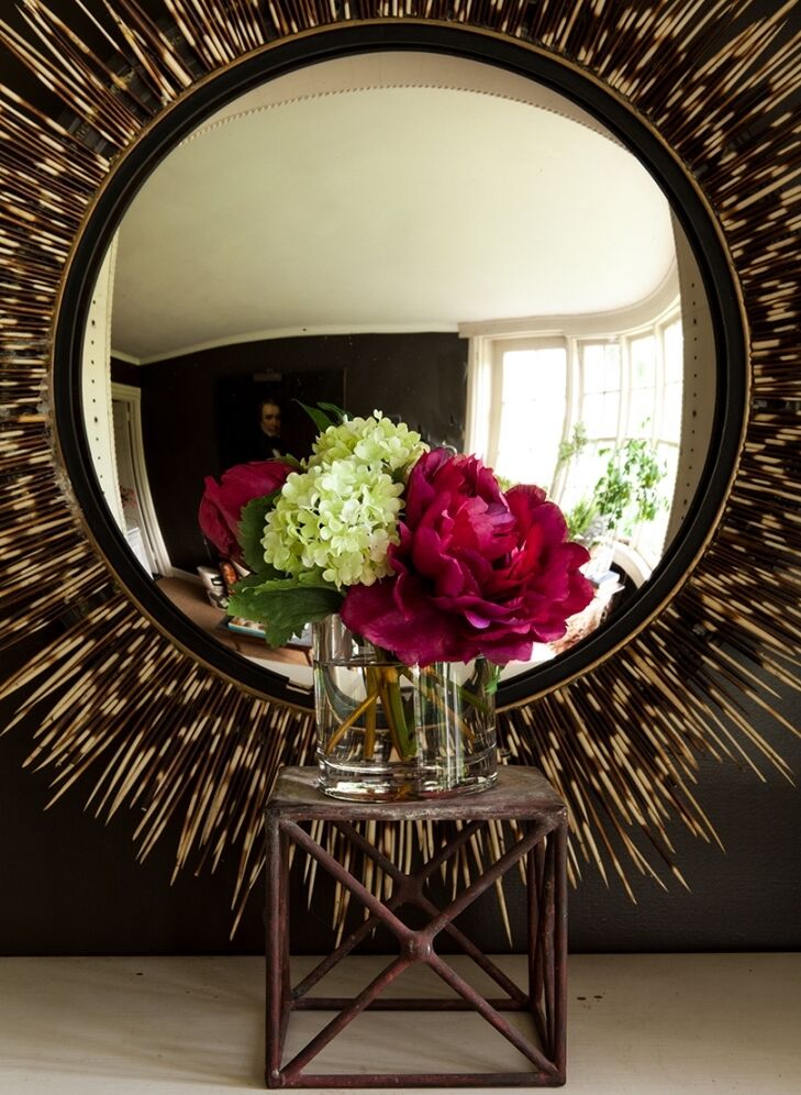 A round mirror with a spiked frame reflects a room with a window. In front of the mirror, a vase with pink and white flowers sits on a small wooden stand.