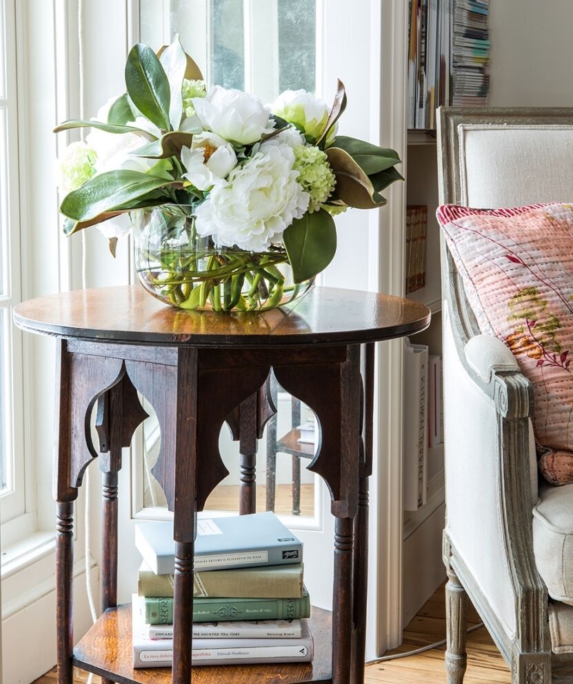 A small wooden side table with an ornate design holds a glass vase filled with white flowers and green leaves. Books are stacked on the lower shelf. The table is next to a cushioned armchair with a patterned pillow, near a window.
