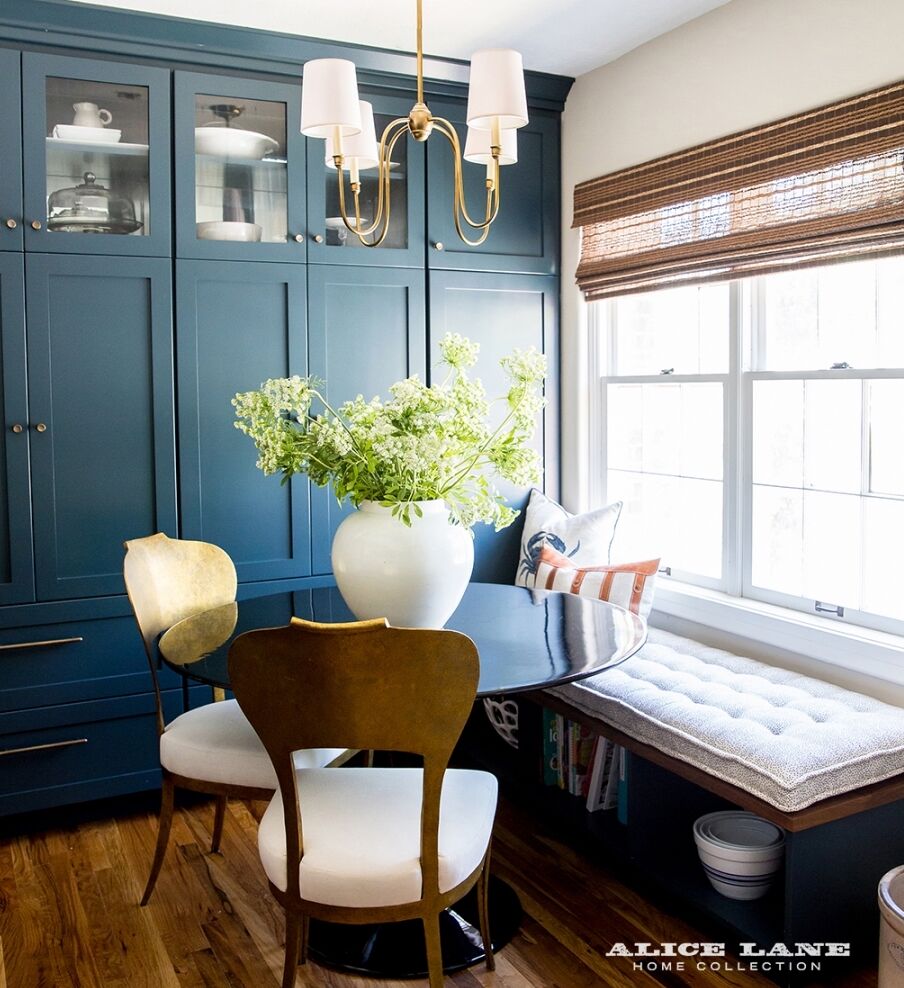 A cozy dining nook features a round black table with a white vase of green flowers. Two elegant chairs, a cushioned window bench with pillows, and navy blue cabinetry with a window above complete the space. A brass chandelier hangs overhead.