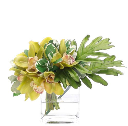 A bouquet of yellow flowers with green leaves in a small, clear square vase. The arrangement includes leafy accents and features vivid, natural colors against a white background.